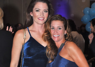 Two women smiling at a party, one in a blue velvet dress with a shoulder bag, the other in a satin blue dress, with a festive background.