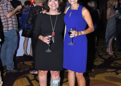 Two women smiling at a social event, one in a black dress and leg brace, the other in a blue dress, both holding wine glasses.
