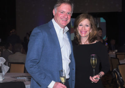 A man and a woman holding champagne glasses at a formal event, smiling at the camera, with event lighting in the background.