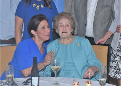 A woman in a blue dress smiles and leans towards an elderly woman in a teal sweater at a table with desserts and drinks.