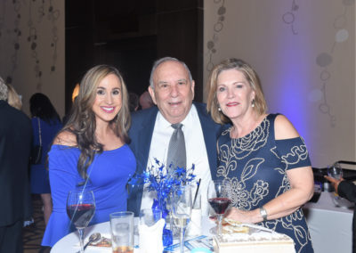 Three people smiling at a formal event, with two women and a man standing around a table with wine glasses and a blue floral centerpiece.