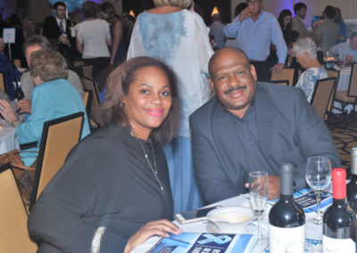 A couple smiling at the camera while seated at a dinner table with wine bottles during a gala event.