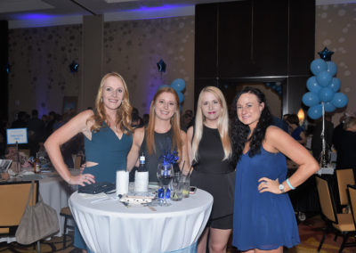 Four women smiling at a gala event, standing behind a table with blue decorations.