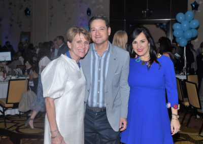 Three people smiling at a formal event with blue balloons and tables in the background.