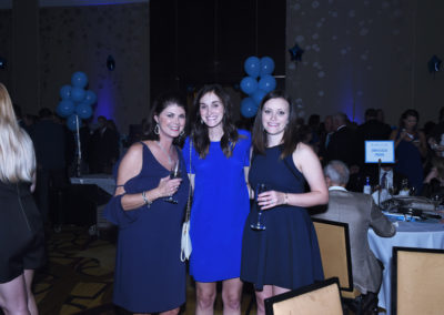 Three women smiling at a formal event with blue balloons, holding wine glasses.