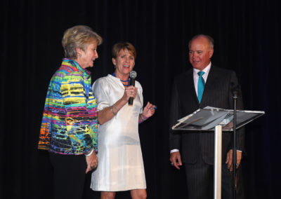 Two women and a man on stage, one woman speaking into a microphone while the others listen. they stand near a podium in a dimly lit room.