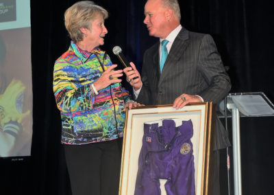 An elderly woman and a man stand next to a framed sports uniform, discussing into microphones at an event.