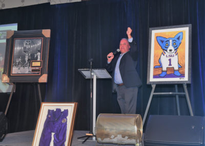 Man in a suit speaking at a podium, gesturing with raised hand, with a painting of a blue dog and other memorabilia displayed at an auction event.