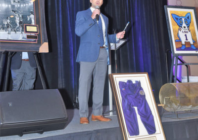 Man in a suit speaking into a microphone on stage at an event, with sports memorabilia displayed around him.