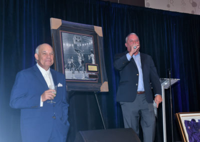 Two men in suits at an event, one holding a microphone speaking and the other holding a drink, with a framed photograph on display beside them.