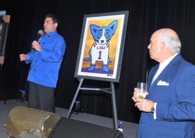 A man in a blue shirt speaks into a microphone at an event while another man in a suit holds a glass, with a painting of a cartoon dog in an lsu jersey between them.