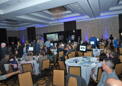 People attending a formal event inside a banquet hall with a presentation screen and scattered seating.