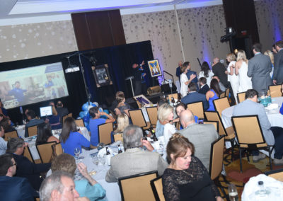 People at a formal fundraising event with dining tables, a stage, and screens displaying images.