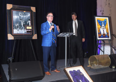 Two men speaking at a podium during an event, flanked by framed artwork and sports memorabilia.