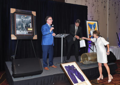 A charity auction event with a man speaking at a podium, another man assisting, and a woman raising her hand to bid, surrounded by displayed artwork.