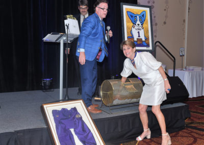 Auctioneer holding a microphone gesturing towards a woman spinning a raffle drum, with framed sports memorabilia displayed onstage.