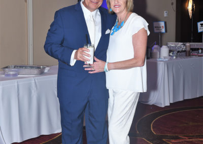 A man and a woman, dressed in formal attire, smiling and standing together at a social event, each holding a drink.