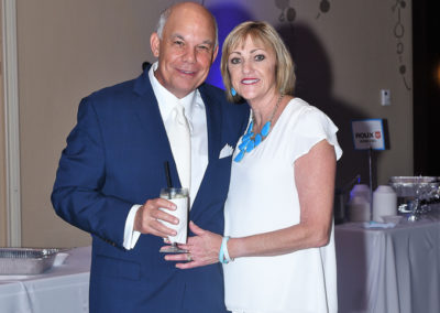 An older man in a navy suit and a woman in a white dress posing together at a formal event, holding a drink.