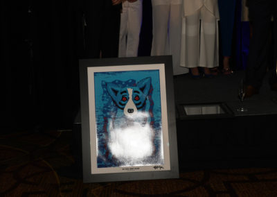 Artwork depicting a blue and black raccoon displayed on an easel at a formal event, with people partially visible in the background.