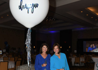 Two women smiling, standing next to a large balloon with "champagne" written on it, surrounded by champagne bottles and glasses at an event.