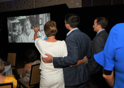 A couple watches a slideshow at an event, with the woman pointing at a photo of an elderly man on the screen.