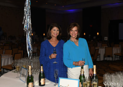 Two women smiling at a gala event, holding wine glasses with tables and champagne bottles in the background.