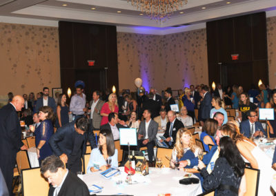 People attending a gala event in a large banquet hall with round tables, guests sitting and mingling, and a blue-lit stage in the background.
