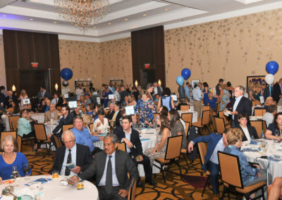 A banquet hall filled with people sitting at tables, decorated with blue and white balloons, during a formal event.