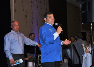 A man in a blue chef's jacket speaks into a microphone at a podium during an event, with another man standing behind him.