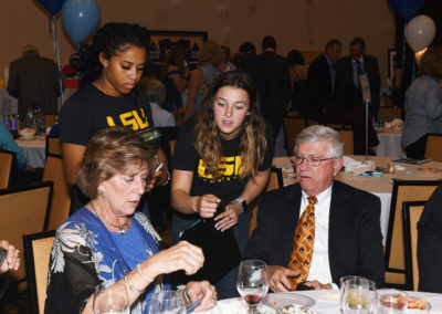 Two young women in lsu shirts talking to an older man and woman seated at a banquet table with blue and white balloons.