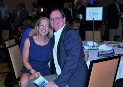 A smiling couple seated at a gala event, the woman in a blue dress and the man in a blazer and glasses.