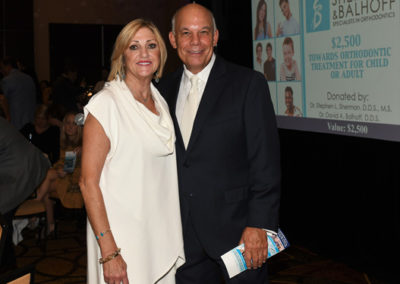 An older couple smiling at a charity event, standing in front of a promotional banner for orthodontic treatment donations.