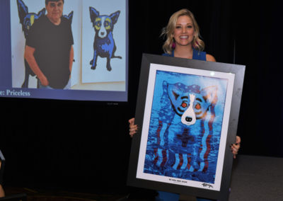 Woman smiling and holding a framed abstract painting of a blue dog, with a projection of a man and similar painting in the background.