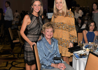 Three generations of women, the eldest seated, posing together at an event with other guests in the background.