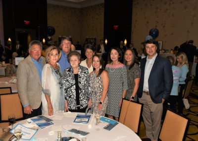 A group of nine adults posing for a photo at a gala event with tables, chairs, and blue balloons in the background.