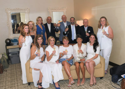 A group of eleven adults in a living room, dressed in white, smiling and toasting with drinks.