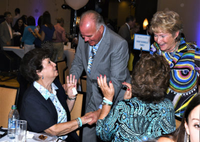 A man greeting three women at a lively event with guests in the background, balloons visible, and everyone appearing engaged in conversation.