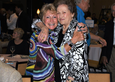 Two elderly women hugging and smiling at a social event with people in the background.