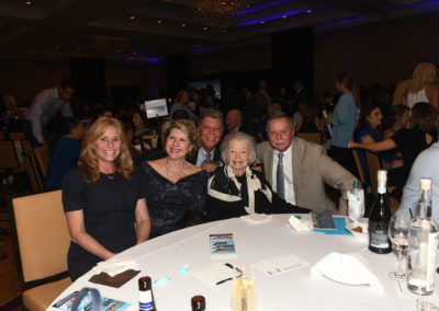 Five adults smiling at a table during a banquet event with other attendees in the background.