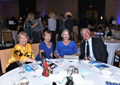 Four older adults smiling at a table during a formal event with people mingling in the background.