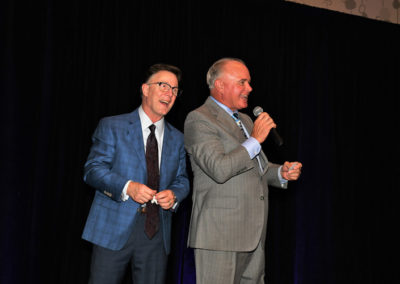 Two men in suits laughing and speaking into a microphone on a stage at an event.