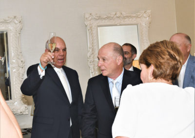 Two men in suits, one raising a glass, standing in a room with ornate mirrors, talking to a woman whose back is facing the camera.