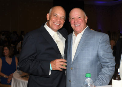 Two smiling men in suits, one in a black suit and the other in a blue checked suit, holding drinks at a social event.