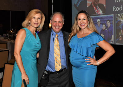 Three people smiling at a formal event; two women in blue dresses flank a man in a suit with a patterned yellow tie. a presentation screen is visible in the background.