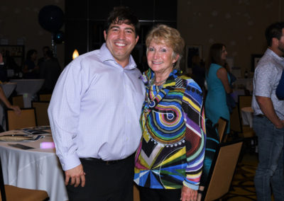 A smiling man and older woman posing together at a gala event, surrounded by tables and other guests in the background.