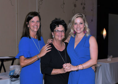 Three women smiling at a formal event, two in matching blue dresses, standing with their hands on the shoulders of the woman in the center.