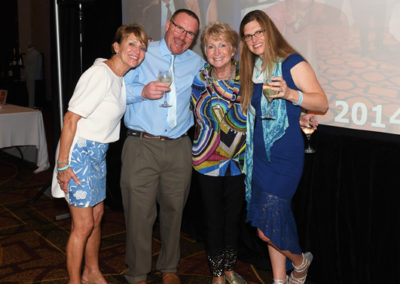 Four cheerful adults posing with drinks at a social event, with a photo projection in the background.