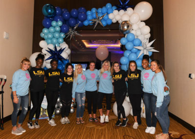 A group of eleven women in lsu t-shirts posing in front of a balloon arch at an indoor event.