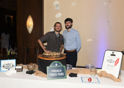 Two men standing behind a food station labeled "french fired fare" at an event, with one holding a wooden tray of appetizers.