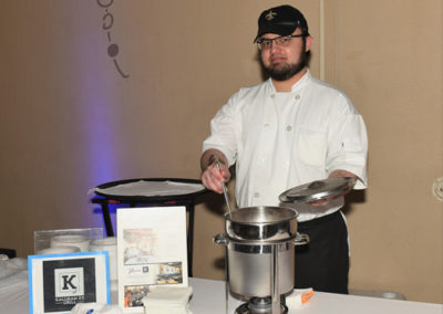 A chef in a white jacket and black cap serving food at a banquet, with decorative items and informational brochures on the table.